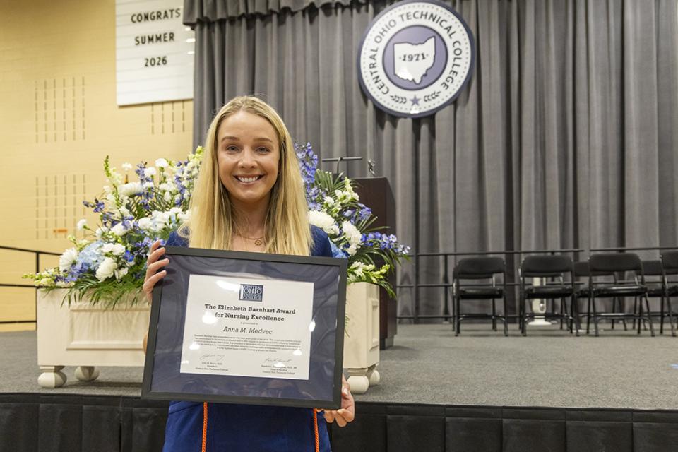 Summer 2026 Barnhart Award recipient Anna Medvec stands in front of the graduation stage holding her framed Barnhart Award certificate.