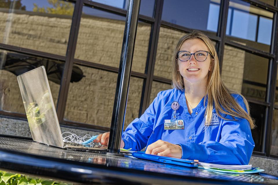 A nursing student in wearing COTC scrubs sits at an outdoor table with a laptop open in front of her.