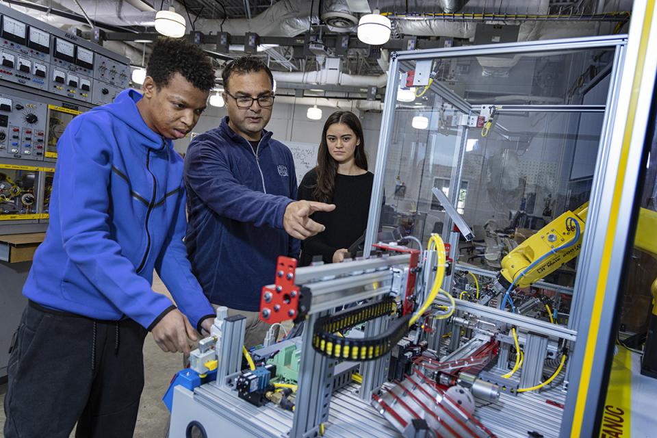 An instructor standing between two students  points to machinery in the engineering lab.