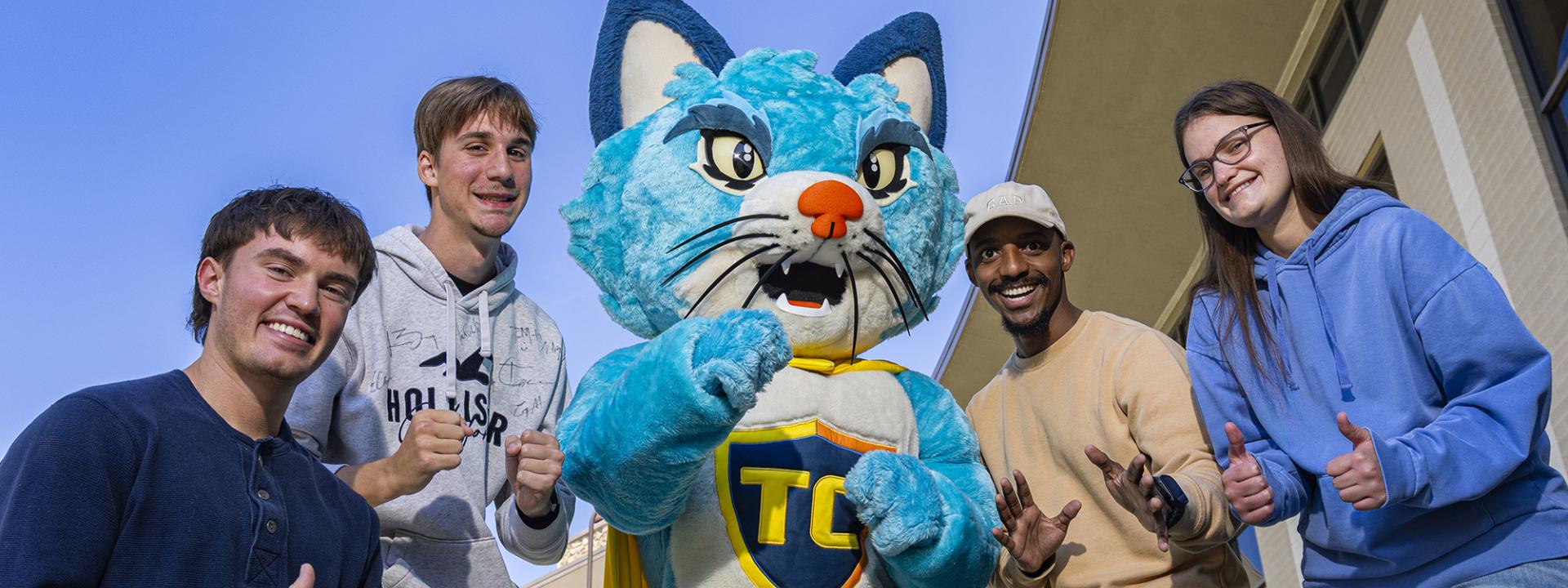 Four students gather around the COTC mascot, TC the cat, making thumbs up gestures with their hands.