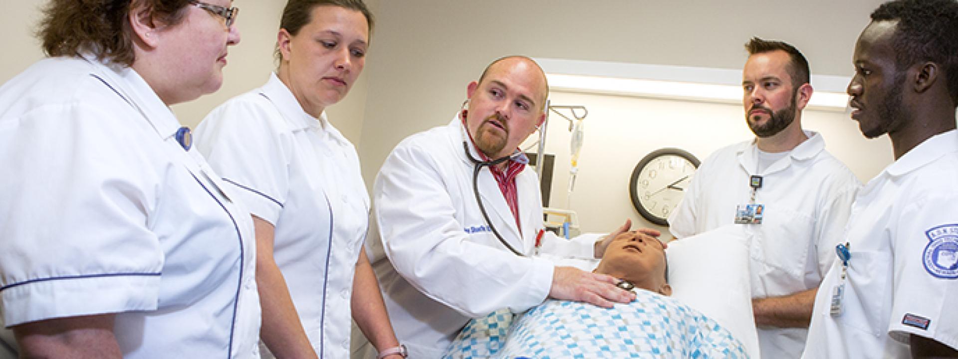 four students standing around professor showing technique on a dummy patient