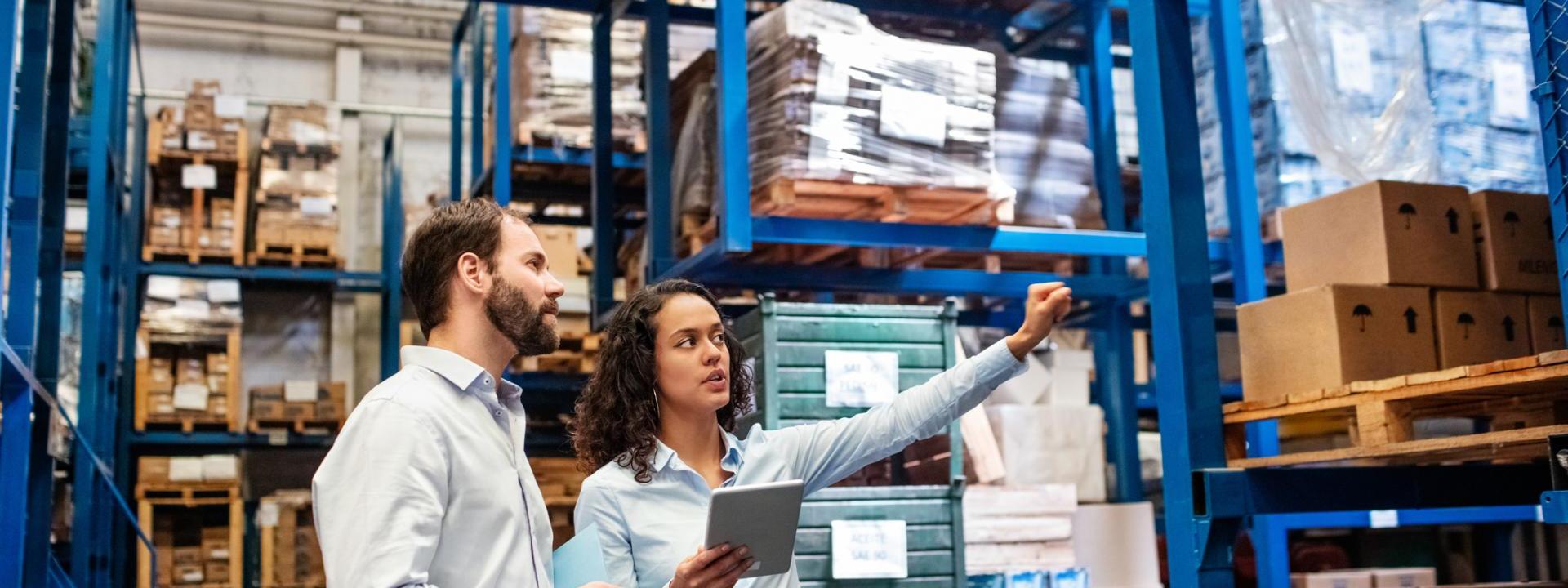 A man and woman look in the direction of her outward stretched hand in a warehouse that has stacks of boxes and product.