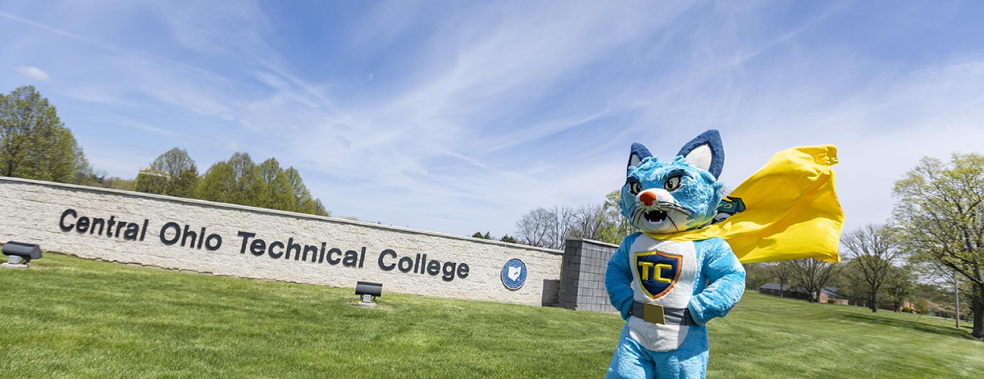 A person in a blue cat  costume with TC on his chest stands on campus with yellow cape blowing in the wind.