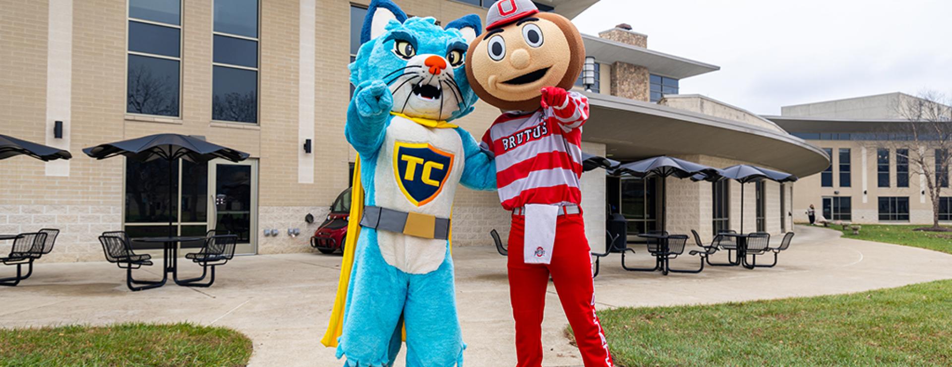 TC the Central Ohio Technical College cat mascot and Brutus Buckeye stand with one arm around each other and the other held out to point a finger toward the viewer.