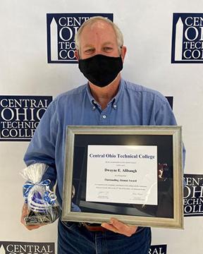 Dwayne Allbaugh wearing blue shirt and mask holding a framed certificate and gift bag