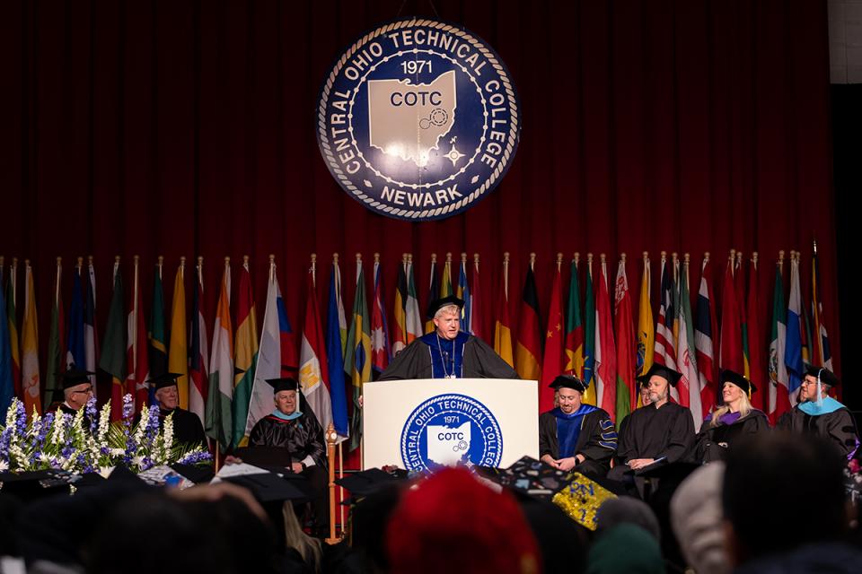 COTC President John Berry stands at a podium dressed in academic regalia overlooking a crowd.