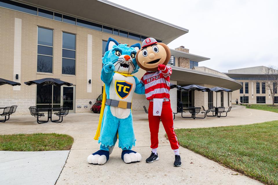 TC the Central Ohio Technical College cat mascot and Brutus Buckeye stand with one arm around each other and the other held out to point a finger toward the viewer.