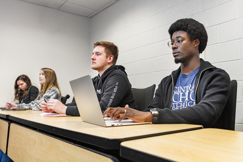 Students sit in a row at a table with one on a laptop looking toward the front of room.