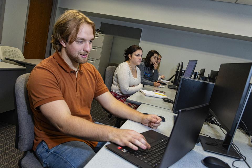 In a row of students, one rests their hands on the keyboard of a laptop computer in a classroom. 