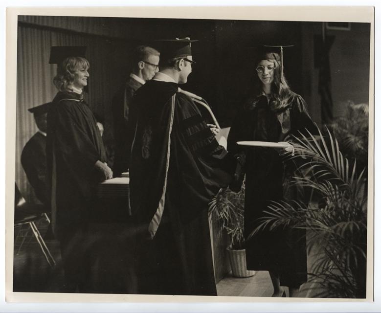 In a black and white photo, Dr. Barnes awards COTC's first associate degree to a student in graduation regalia on state at a commencement ceremony while faculty stand on stage in the background.