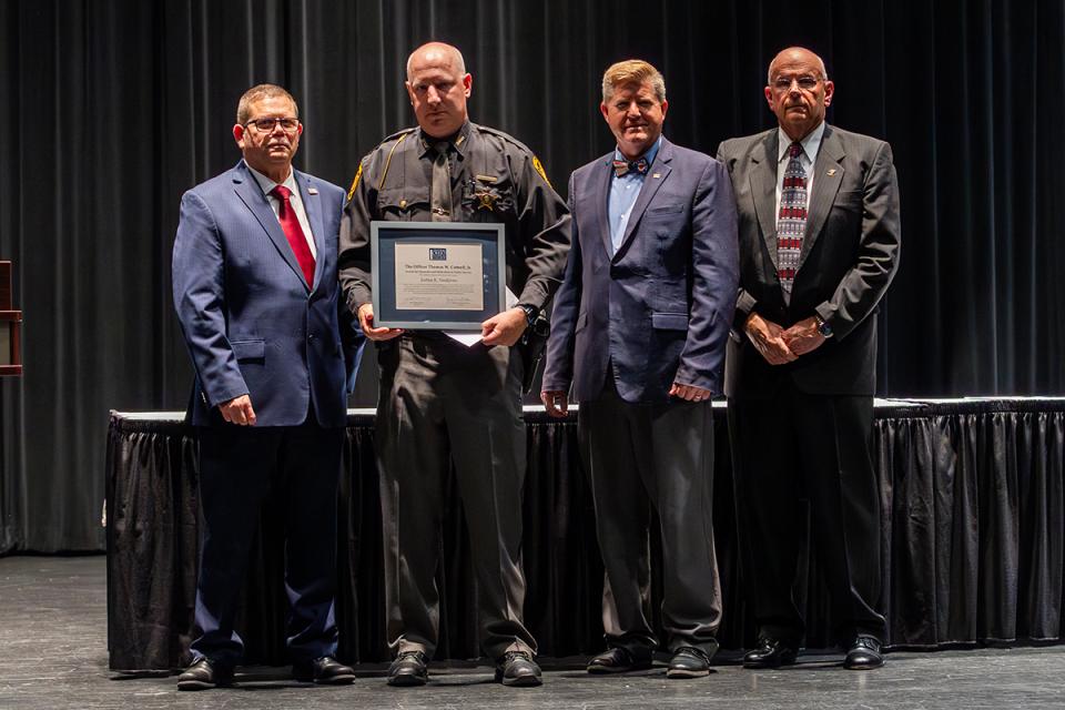 Joshua Snodgrass stands on stage holding his award certificate with Commander James Rease; COTC President John Berry, PhD;  and Commander Jeff Sowards.