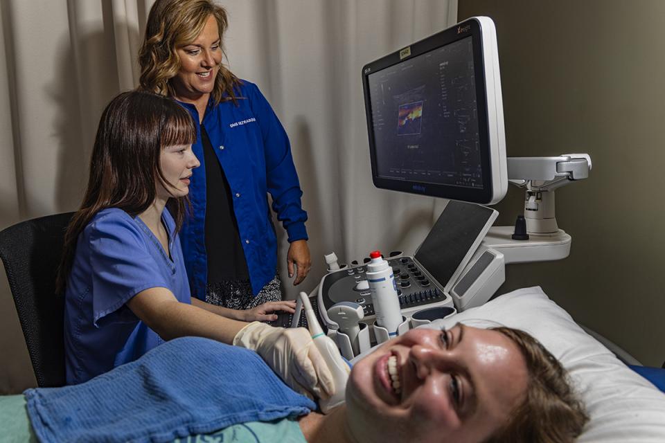 A student practices using an ultrasound machine on another student while an instructor observes.