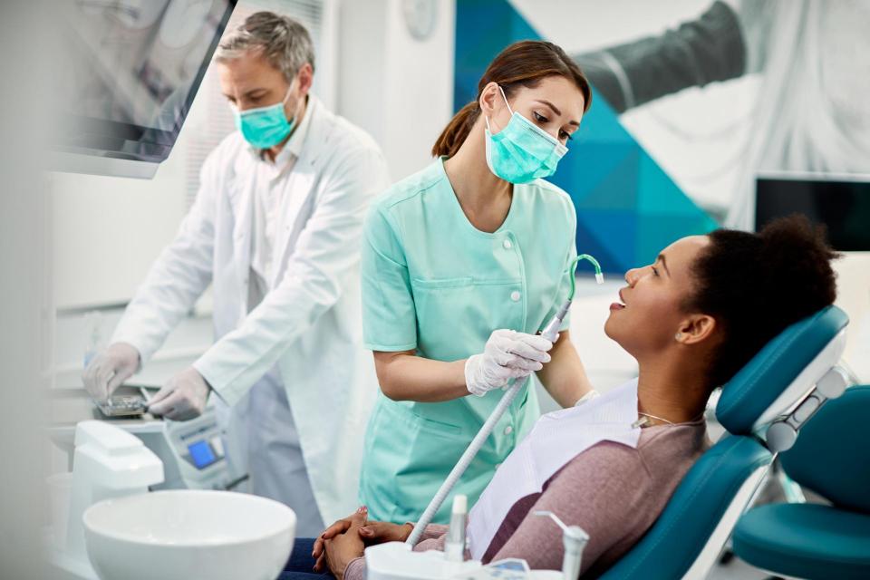 Dental assistant holds a saliva ejector while looking at a patient's mouth with a dentist in the background. 