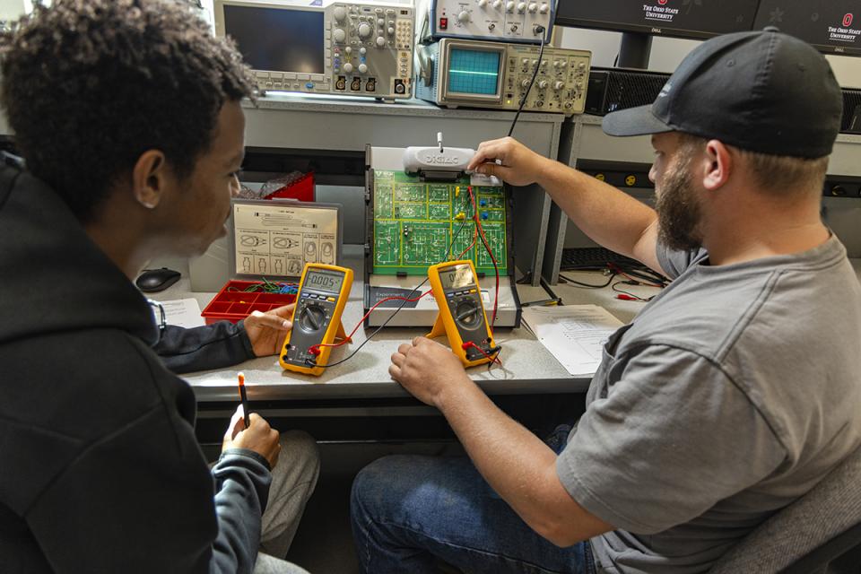 Students sit at a machine taking electrical readings from a handheld device.