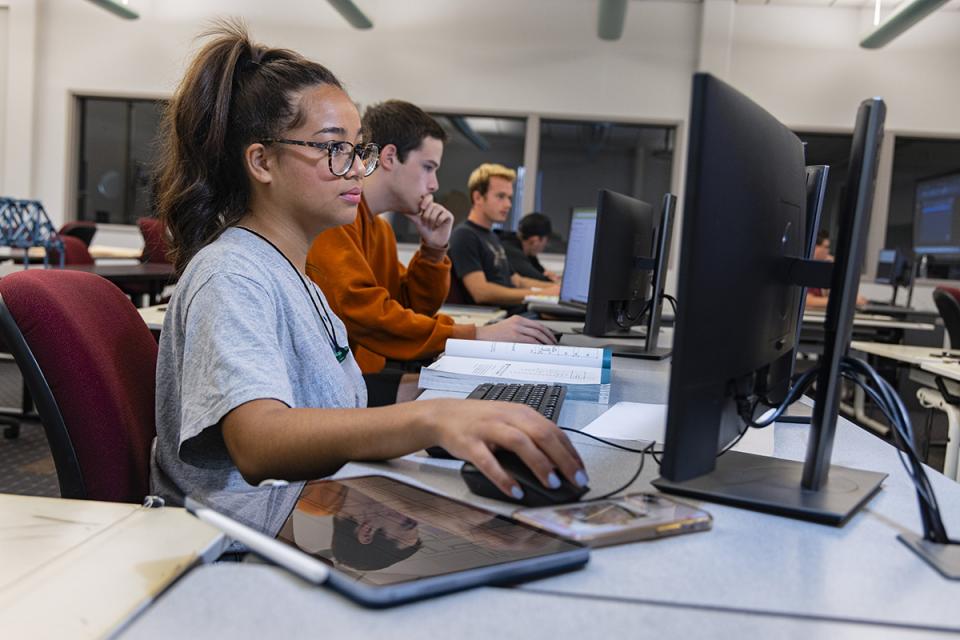 A student seated at the end of a row of students looks at a computer monitor with her hand on a mouse.