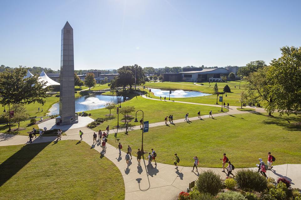 A view from the rooftop of students walk on the campus sidewalk with the bell tower in the foreground and ponds and academic buildings in the background.