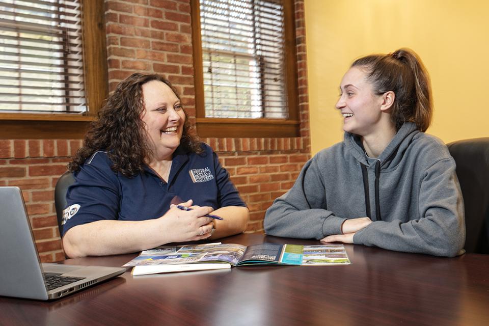 A staff member sits next to a student in conversation with promotional materials and a laptop on the table.