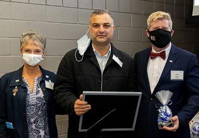 Short-haired woman wearing a mask, man with mustache holding framed certificate and man in suit wearing a mask