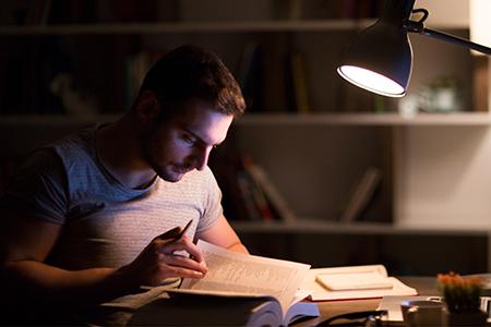 A male students studies from a book with a desk lamp on at home. 