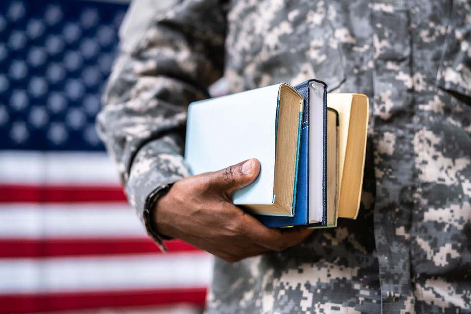 Close-up photograph of a man in military uniform holding books in front of an American flag.