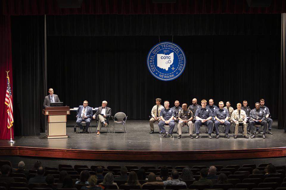The stage of the May 21, 2026 Peace Officer Basic Training program graduation at COTC includes the graduates and State Representative Kevin Miller speaking at the podium.