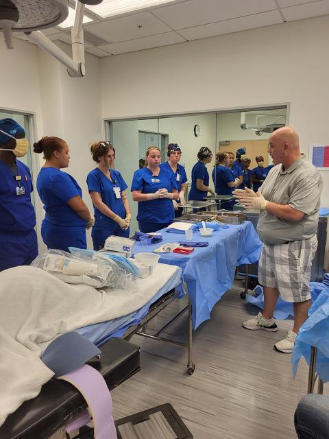 Students in scrub stand next to a table of instruments while an instructor gives instruction from the other side.