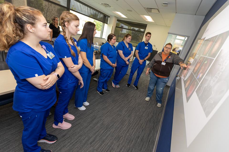 Students stand in front of a white board while a instructor points to information on the board.