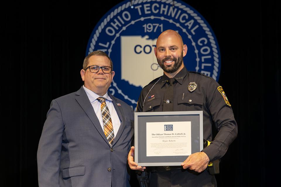 Skyler Roberts holds a framed award certificate on stage next to Peace Office Basic Training Commander James Reese. 