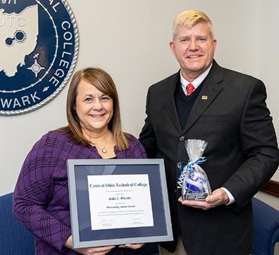 dark haired woman wearing purple sweater holding framed certificate standing with blonde man in suit