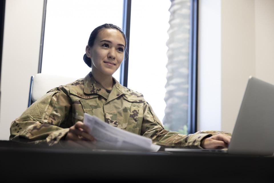 A woman in an Air Force camouflage uniforms is seated at a desk with one hand resting on an open laptop and the other holding paper.