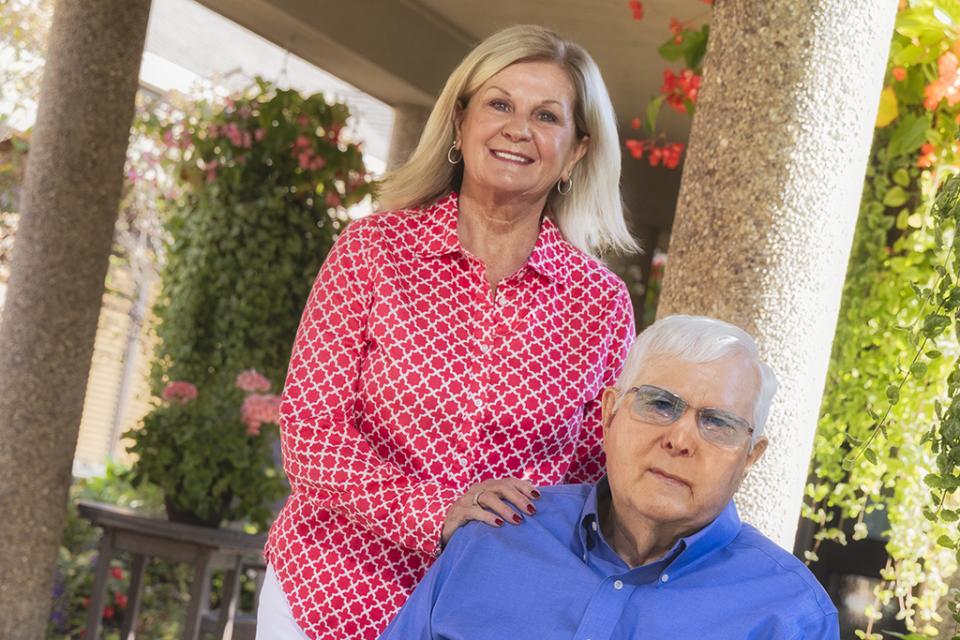 Portrait of Bev Donaldson and a man sitting outside on a sunny day at a facility adorned with plants.