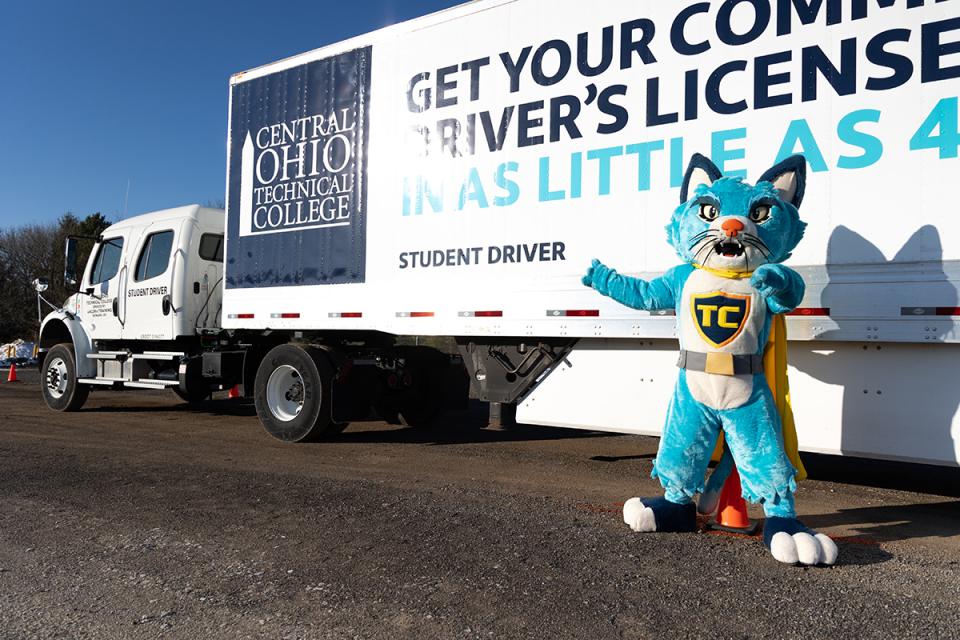 A blue cat mascot stands in front of a semi truck with the COTC logo on  it.