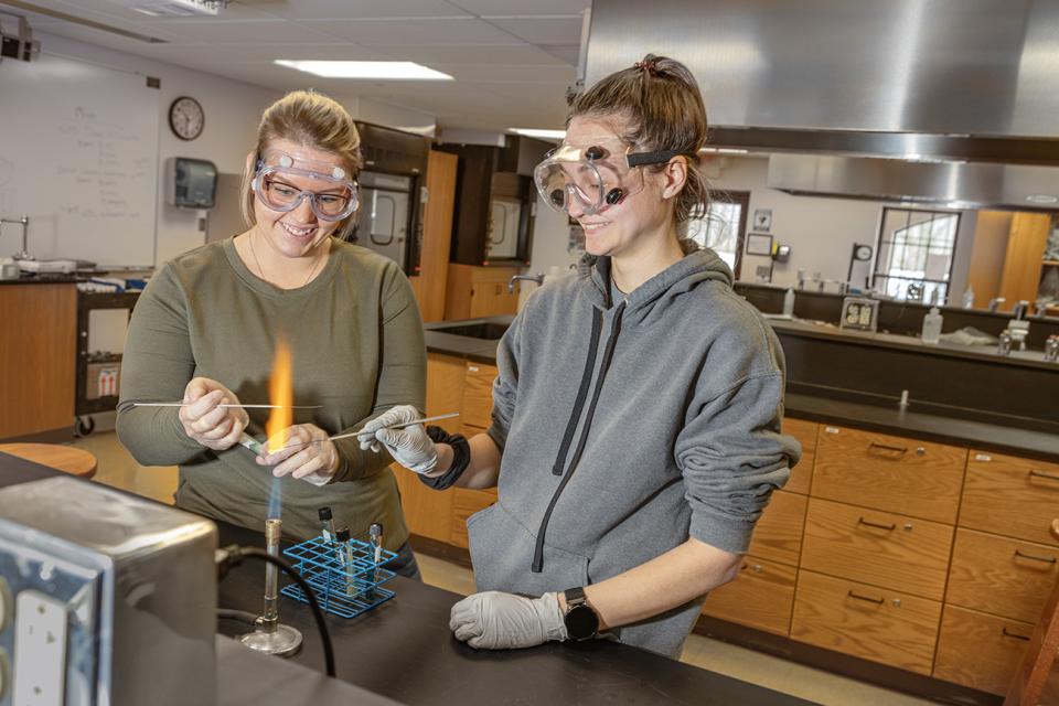 Two students wearing safety goggles do classwork with a bunsen burner. 