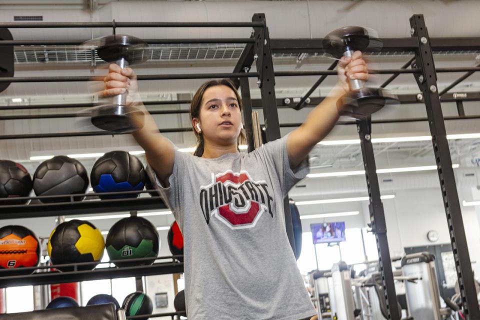 A student in a weight room holds dumbbells in her hands outstretched  in front of her in motion. 