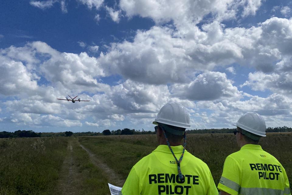 Two men in safety vests that say "remote pilot" stand in a field operating a drone.