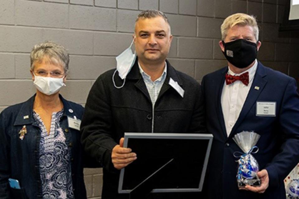 Short-haired woman wearing a mask, man with mustache holding framed certificate and man in suit wearing a mask