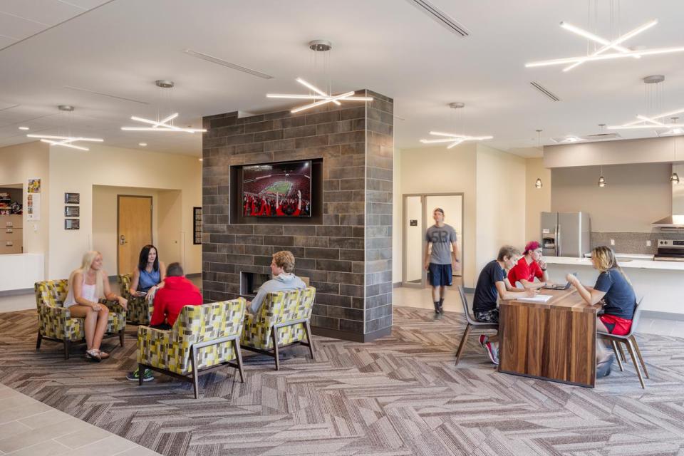 A group of students sits around a fireplace with a mounted tv while another group sits at a table near a kitchenette.