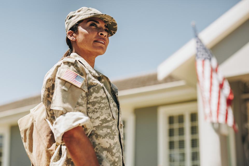 A person in camouflage military attire stands in front of a building  with an American flag displayed.