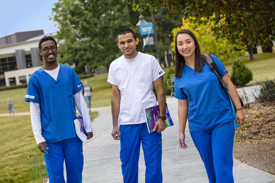 Three students wearing nursing scrubs walk on campus holding their books. 