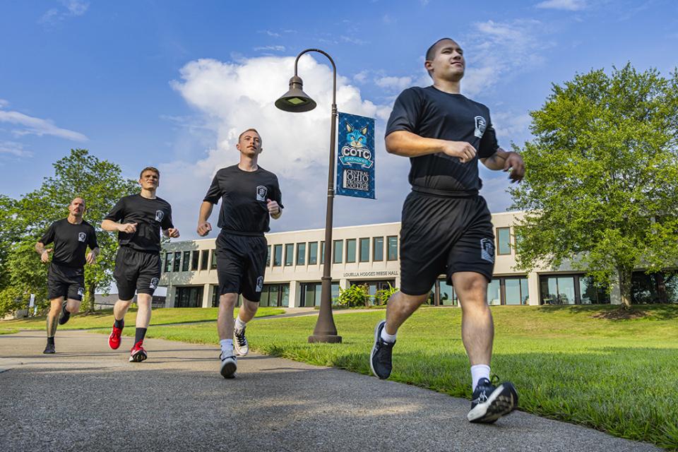 Four cadets in police academy t-shirts and shorts run in a single-file line along the sidewalk on the COTC Newark campus.