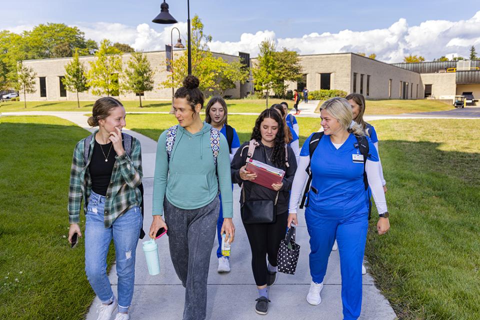 A group of students, some wearing nursing scrubs and some in plain clothes,  walks on the sidewalk between buildings on campus. 