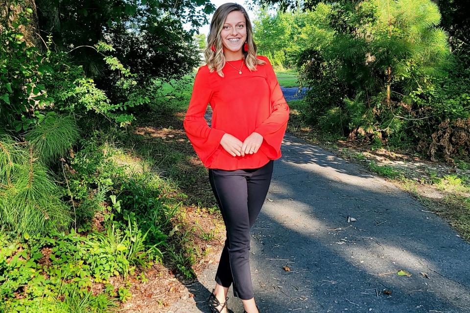 young Caucasian woman with blonde hair and blue eyes in red shirt, black pants, and black sandals poses for a picture