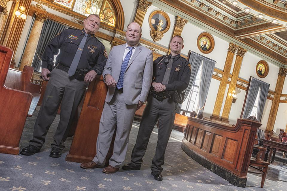 Two men in police uniforms and one man in a suit stand inside a courtroom of the Licking County Courthouse.
