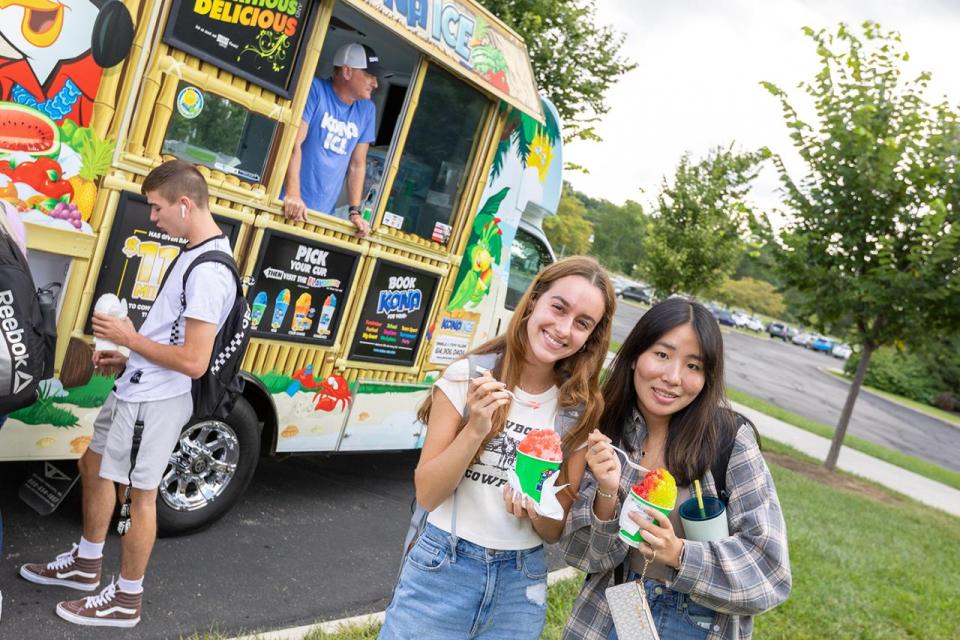Two smiling female students stop for a portrait with their Kona Ice as other students line up at the truck behind them during Welcome Week.