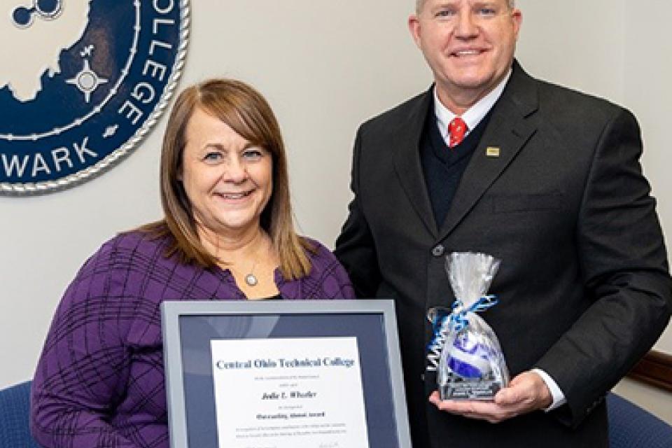 dark haired woman wearing purple sweater holding framed certificate standing with blonde man in suit