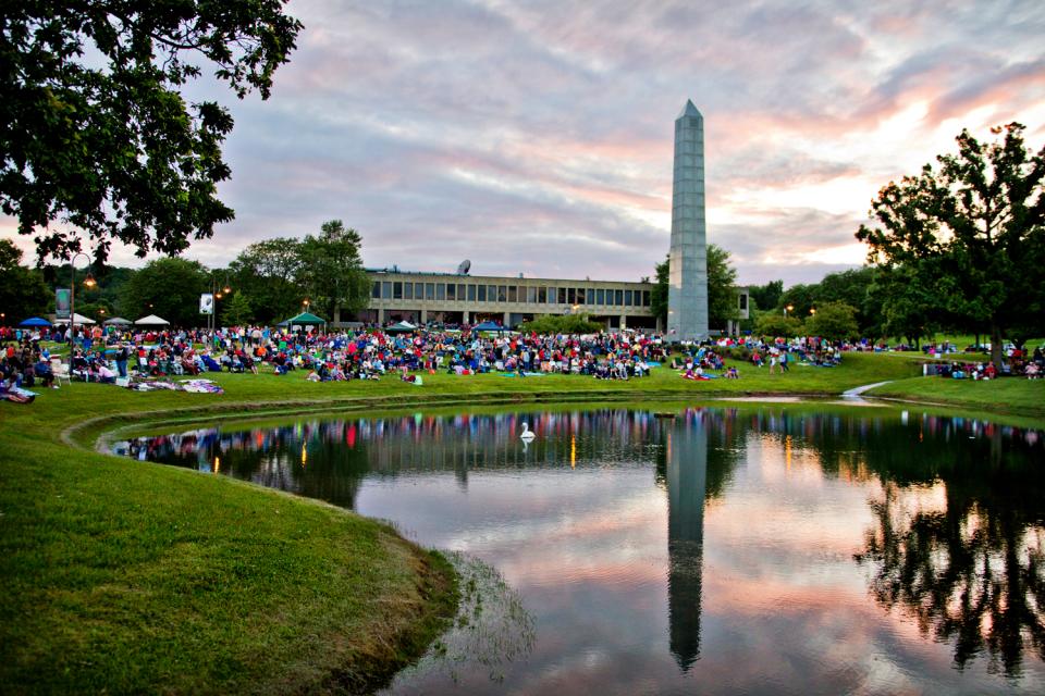 A crowd of people sitting on blankets, in chairs or under tents on the lawn of Founders Hall at dusk. 