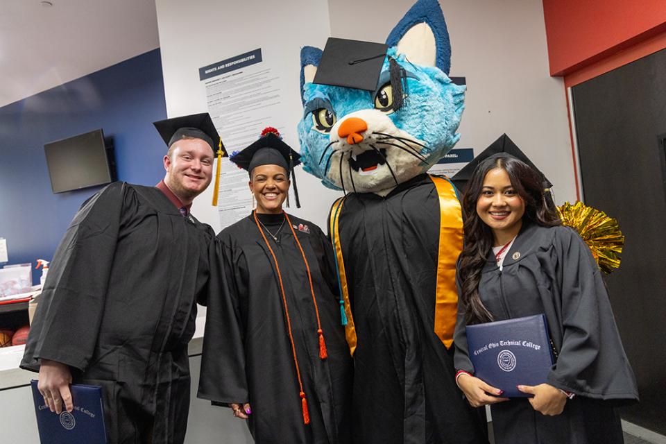 Graduates in their caps and gowns stand with COTC mascot TC the cat.