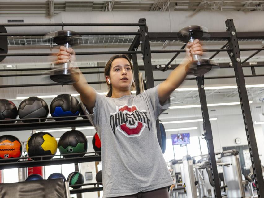A student in a weight room holds dumbbells in her hands outstretched in front of her in motion.