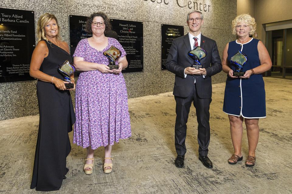 Kim Goudy, Rhonda Carter, Matt Smith representing Ohio State Newark, and Misty Amacher stand in a line holding their Legends of Loyalty Award trophies at a formal event to recognize the recipients. 
