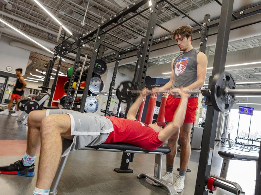 A student uses a bench press while another spots him.